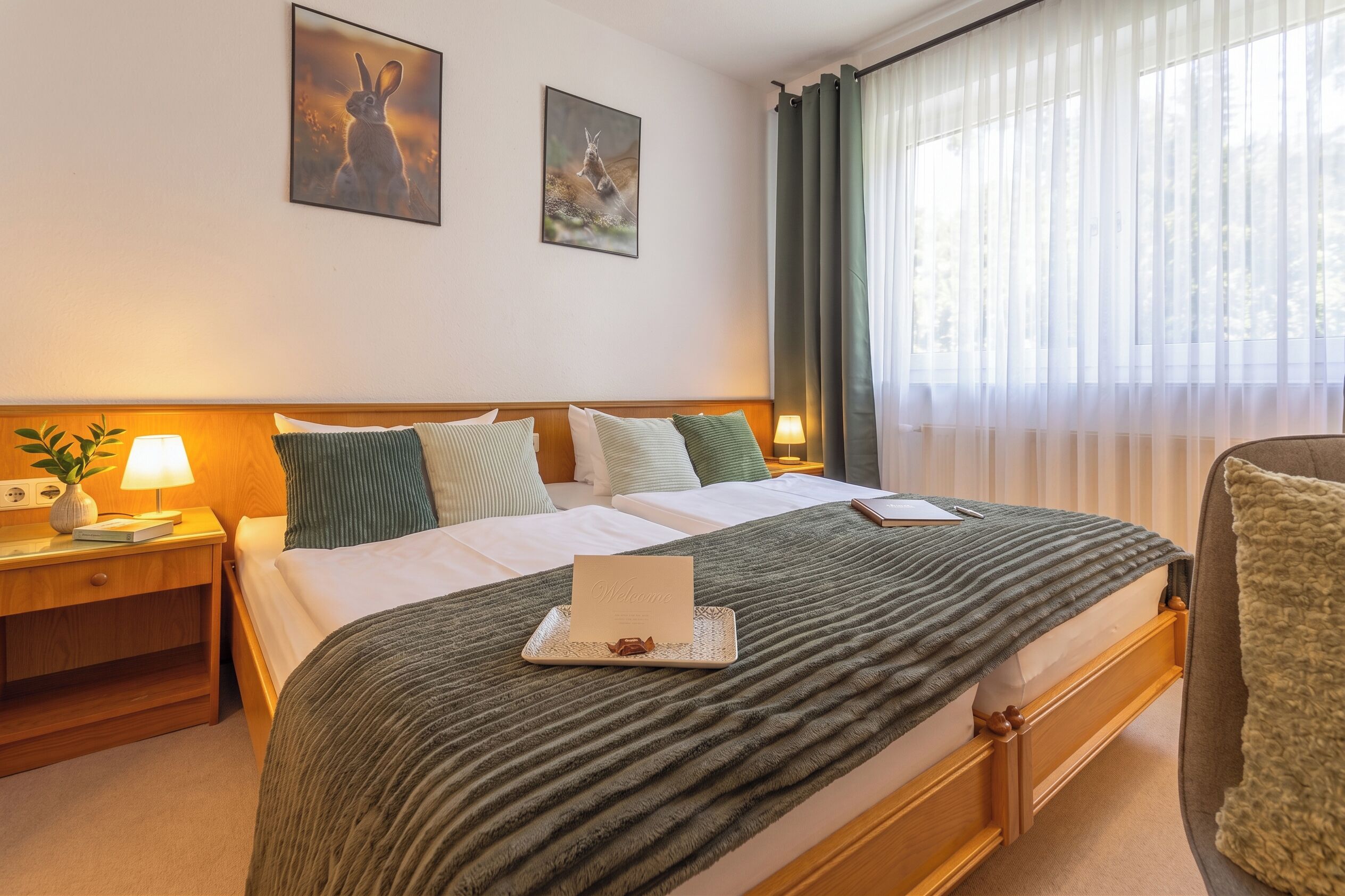 A neatly made bed with a welcome card, a bedside lamp, and a framed picture on the wall.