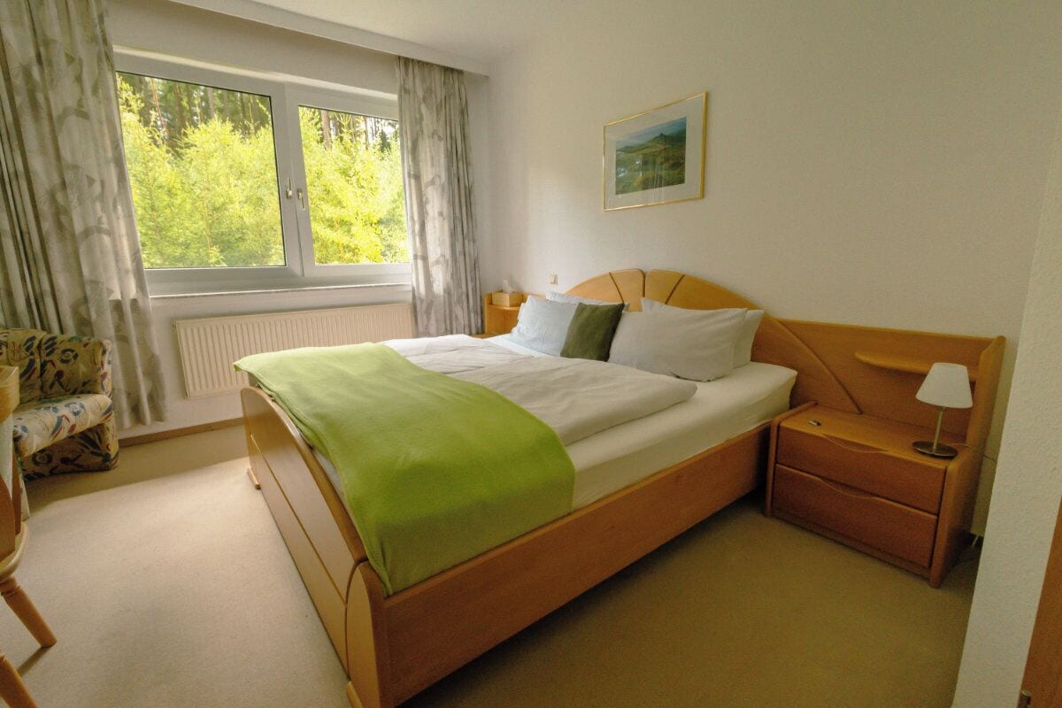 A bedroom with a bed, a nightstand, a chair, a window with curtains, and a painting on the wall.