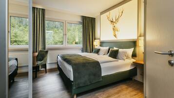 Ein Hotelzimmer mit einem Bett, einem Schreibtisch, einem Stuhl, einem Fenster mit Vorhängen und einem an der Wand hängenden Hirschbild.