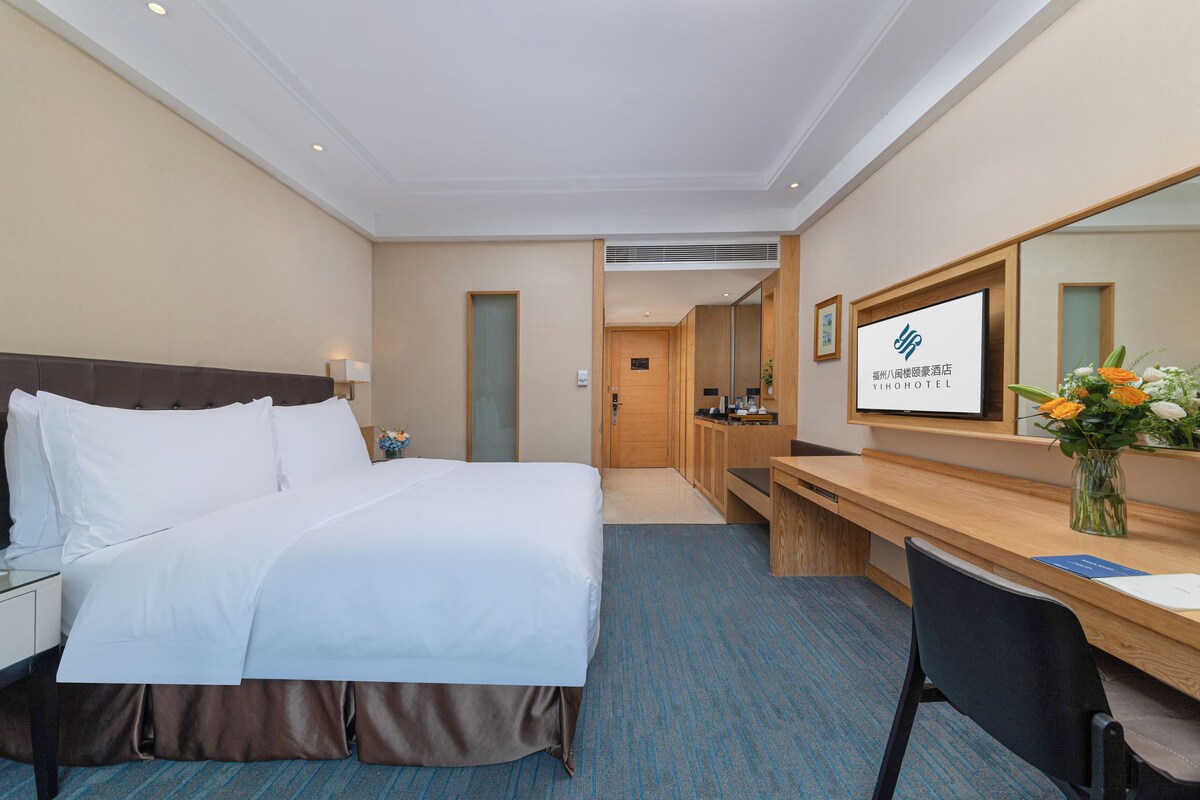A hotel room with a bed, a desk, a chair, a mirror, and a wall-mounted TV.