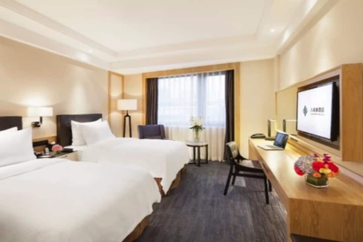 A hotel room with two beds, a desk with a laptop, a TV, and a small table with flowers.