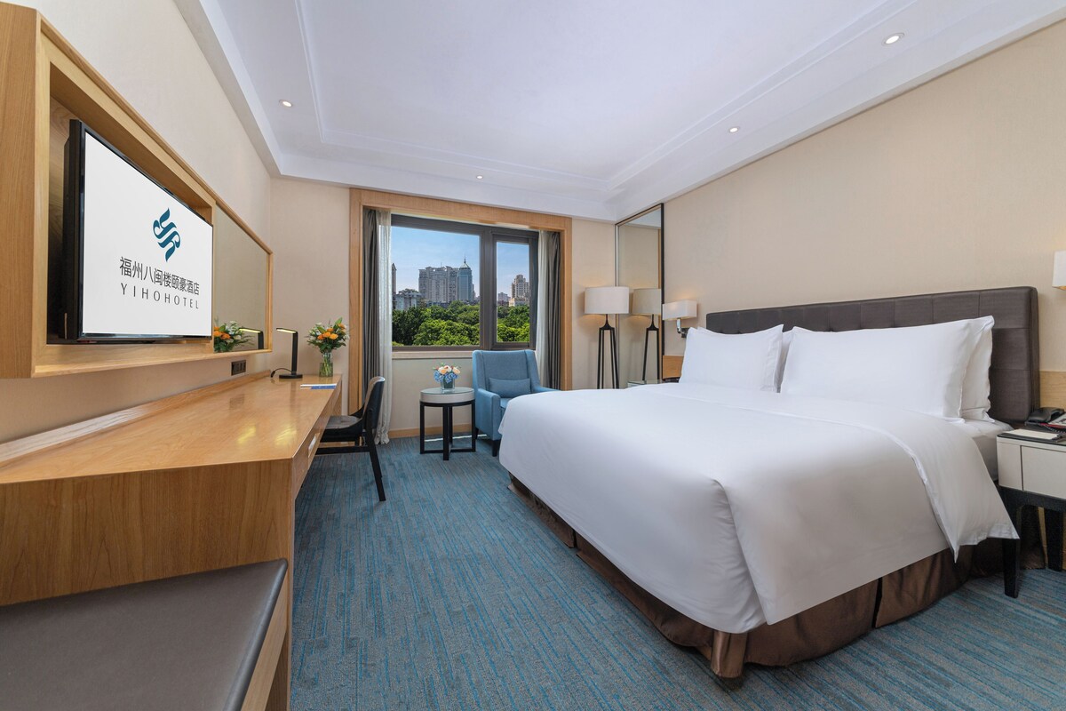 A hotel room with a bed, a desk, a chair, a small table with a vase of flowers, and a large window with a city view.