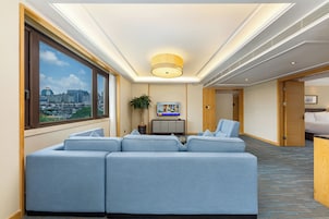 Habitación de hotel con un ventanal grande, zona de estar, televisor y vistas a la ciudad.