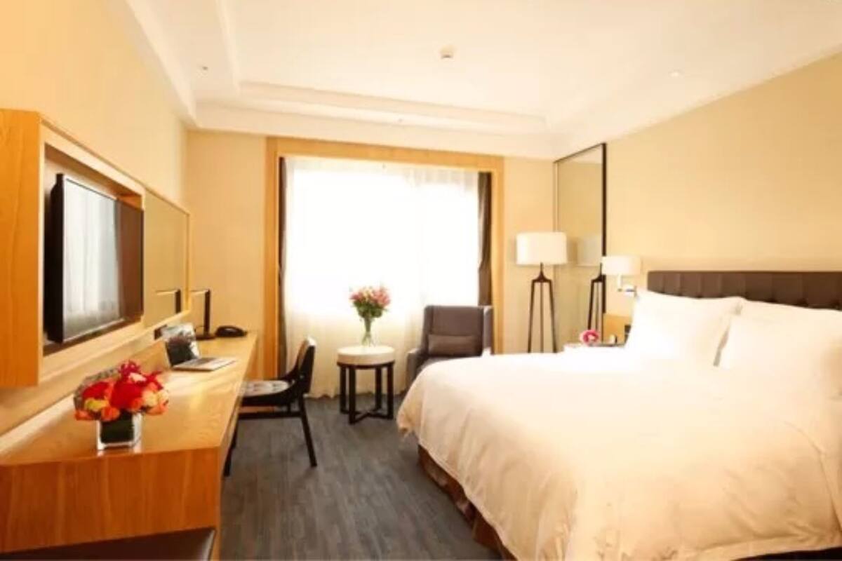 A hotel room with a bed, a desk with a chair, a television, a small table with a vase of flowers, and a lamp.