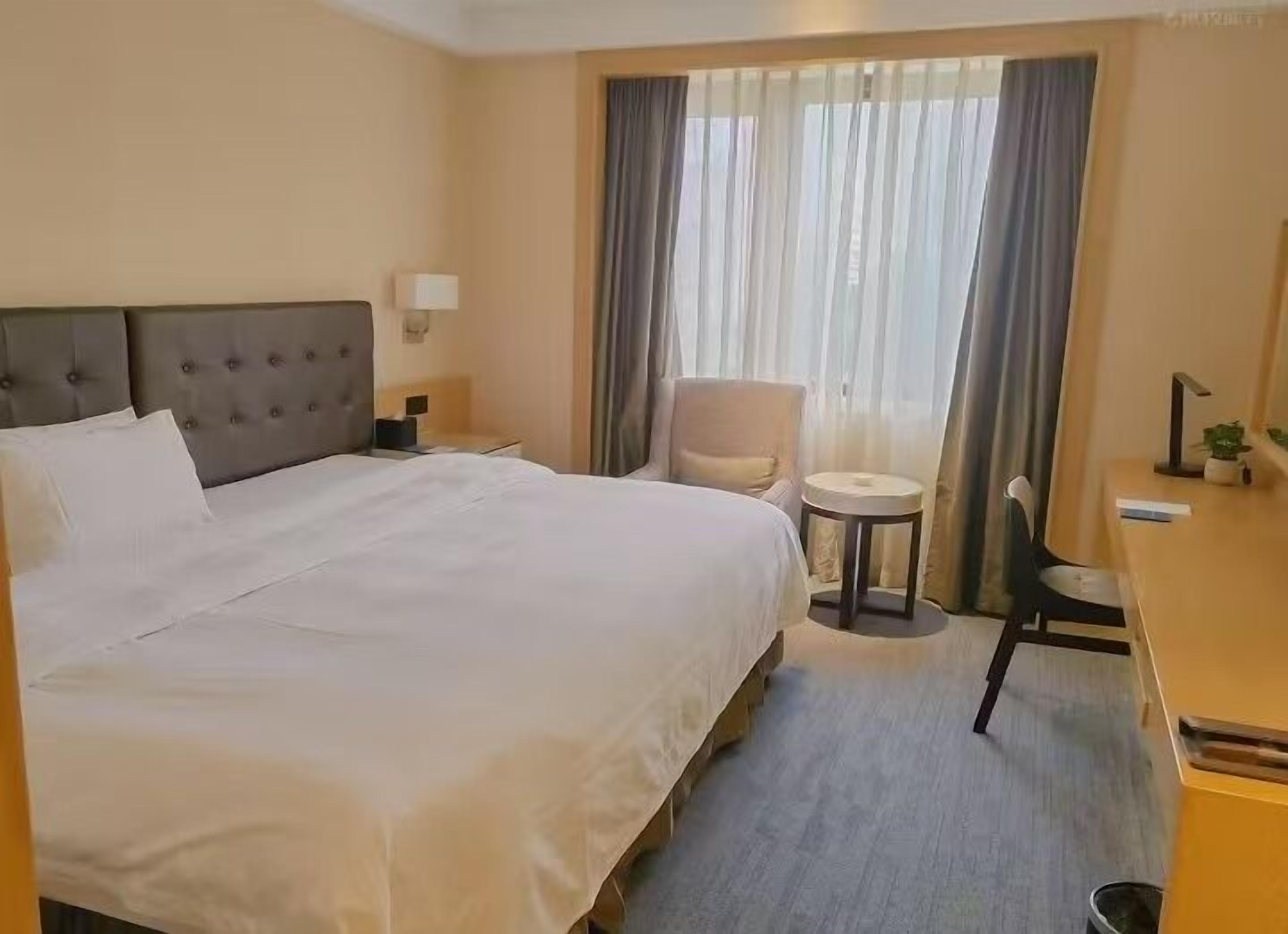 A hotel room with a large bed, a desk, a chair, a small table, and a window with curtains.