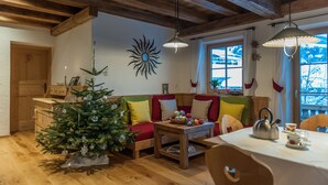 A cozy living room with a wooden ceiling, a Christmas tree, a sofa, a table with a kettle, and a window with curtains.