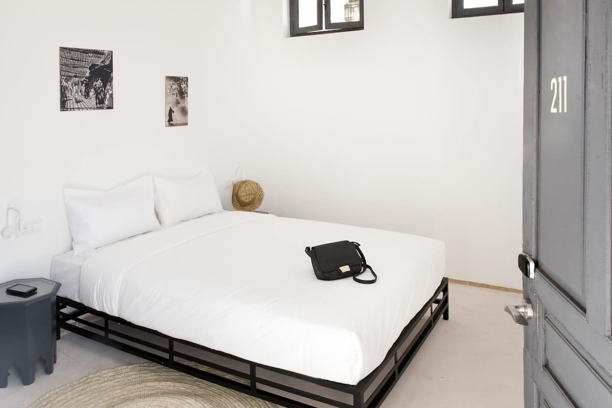 Una cama bien hecha con ropa blanca, un bolso negro sobre la cama, una mesita de noche pequeña y un cuadro colgado en la pared.