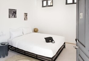 Un letto rifatto con lenzuola bianche, una borsa nera sopra il letto, un comodino e un quadro appeso al muro.
