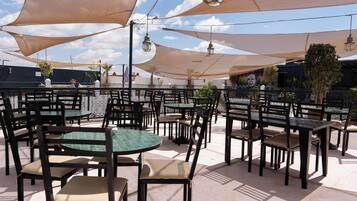 Un patio al aire libre con mesas y sillas redondas, con grandes sombrillas y luces colgantes de estilo farol.