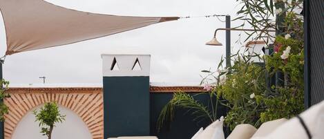Un'area salotto all'aperto illuminata dal sole, con cuscini color crema, una pianta in vaso e un punto luce fissato al muro.
