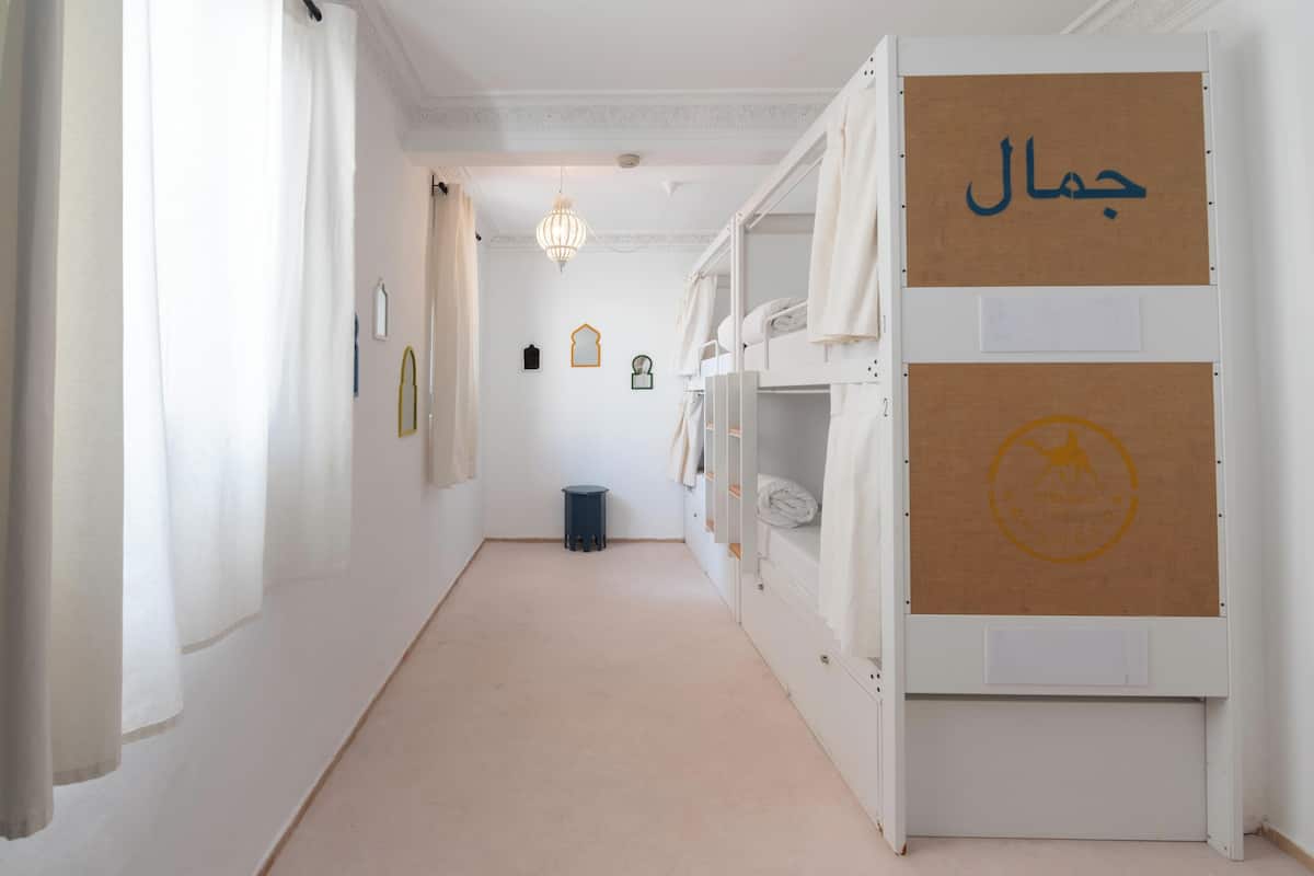 Un pasillo estrecho con literas a un lado, un cubo de basura pequeño y cuadros en la pared.