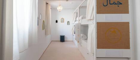 Un couloir étroit, avec des lits superposés d’un côté, une petite poubelle et des tableaux encadrés accrochés au mur.