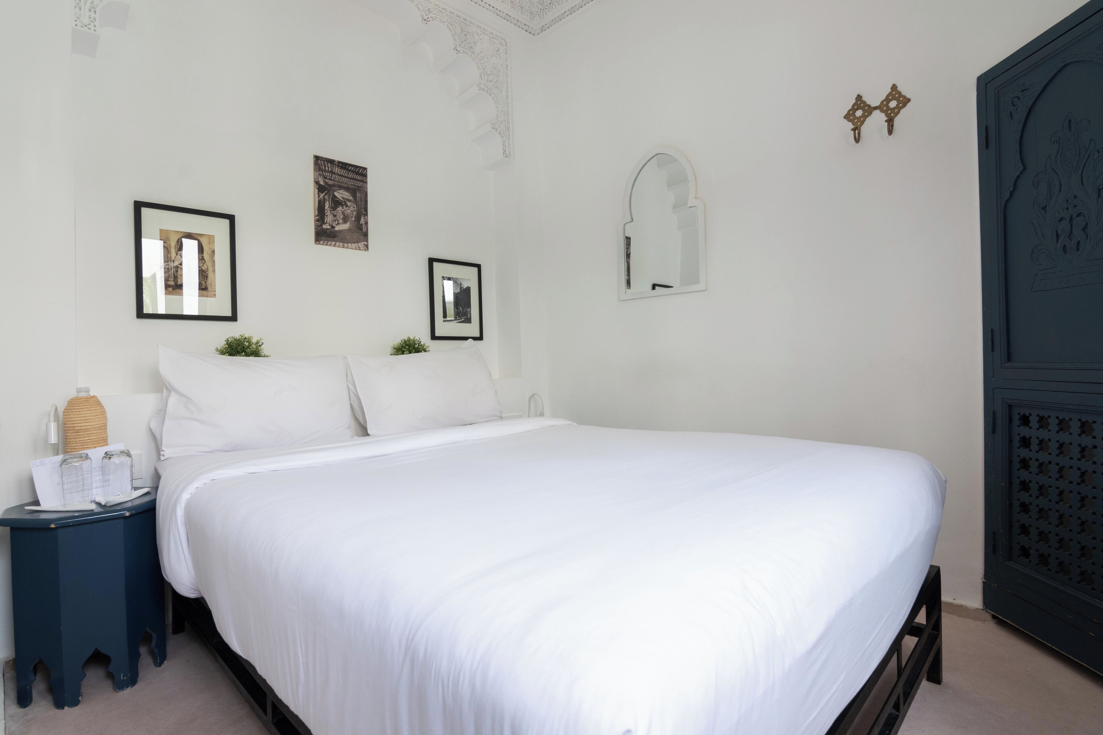 Una cama bien hecha con sábanas blancas, una mesita de noche con una lámpara y cuadros en la pared.