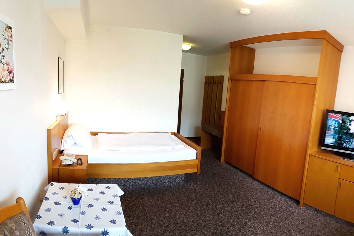 Una camera d'albergo compatta con un letto, un tavolino ricoperto da una tovaglia floreale, una televisione e un armadio in legno.