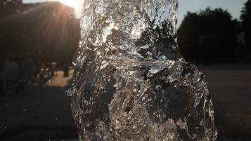 A close-up of a water feature with a reflective surface, possibly a fountain or sculpture, with sunlight creating a lens flare effect in the background.