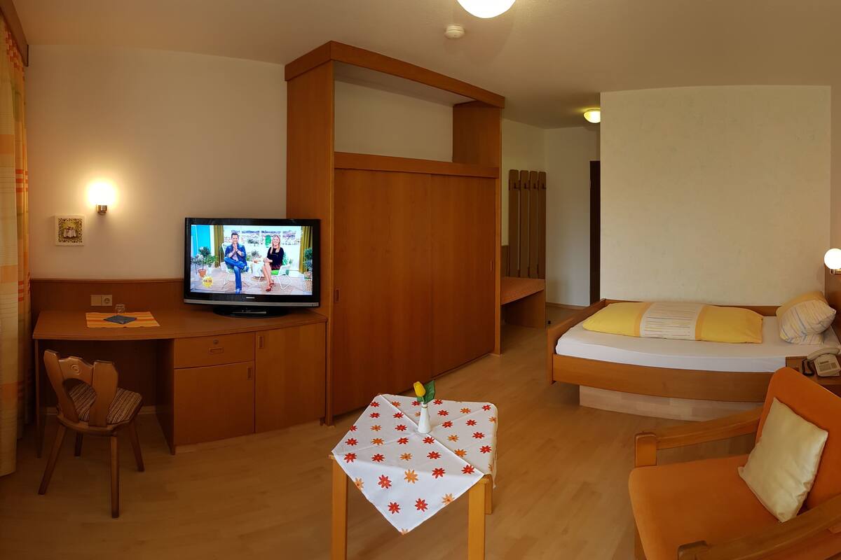 Camera d'albergo compatta con un letto, una scrivania, una televisione e un piccolo tavolo con tovaglia a fiori.