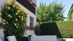 An outdoor seating area with a sofa and planters, a hedge, and a building with flower boxes. - Kurhotel Anders (Bad Fuessing)