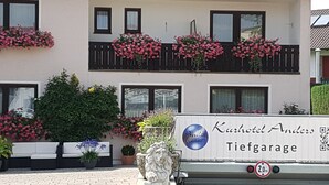 A hotel facade with balconies adorned with pink flowers, a sign reading "Kurhotel Anders Tiefgarage," and a statue in the foreground. - Kurhotel Anders (Bad Fuessing)