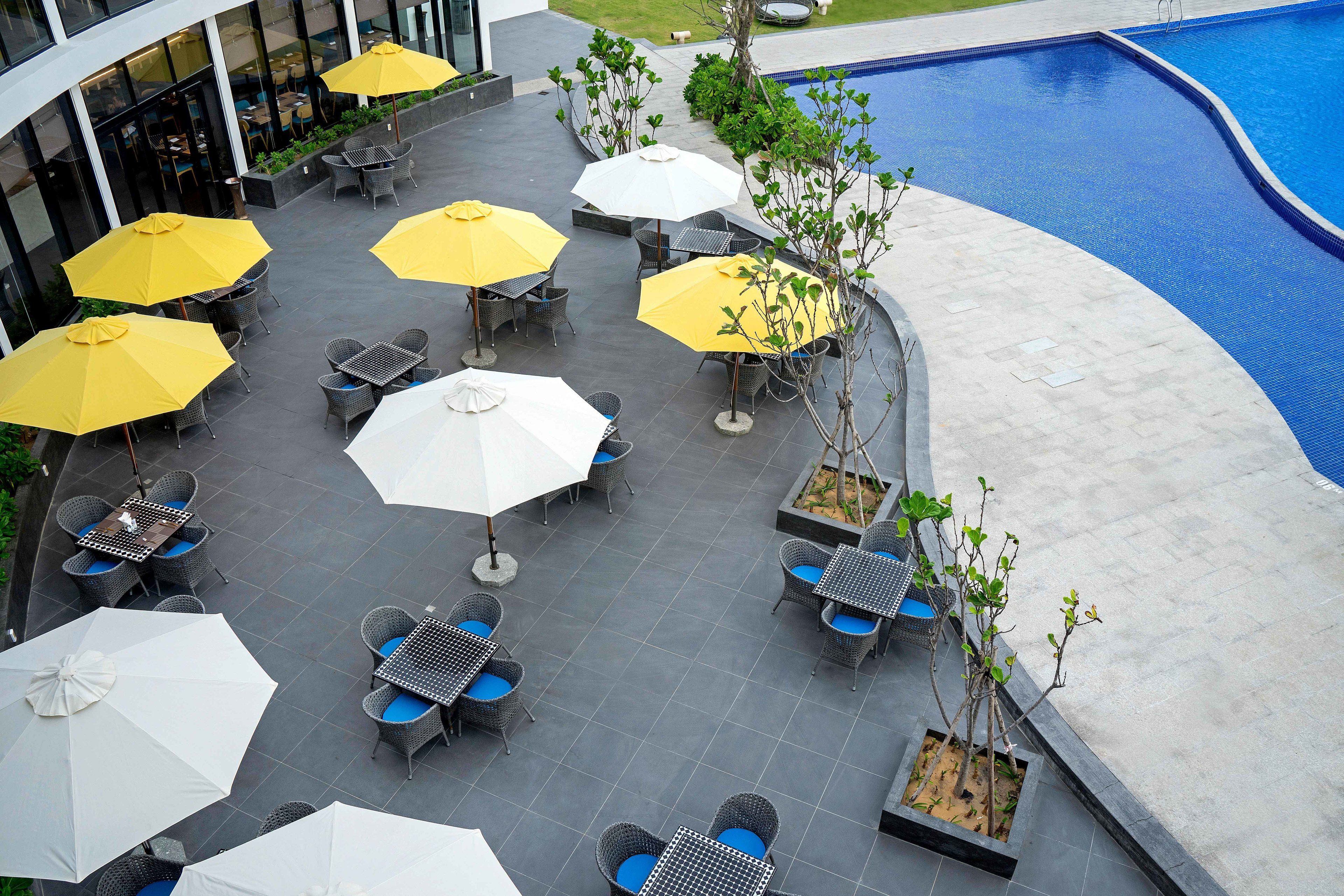 An outdoor patio area with tables and chairs, some with umbrellas, and a pool in the background.