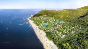 Vista aérea de un complejo turístico frente al mar, con playa, exuberante vegetación y un faro.