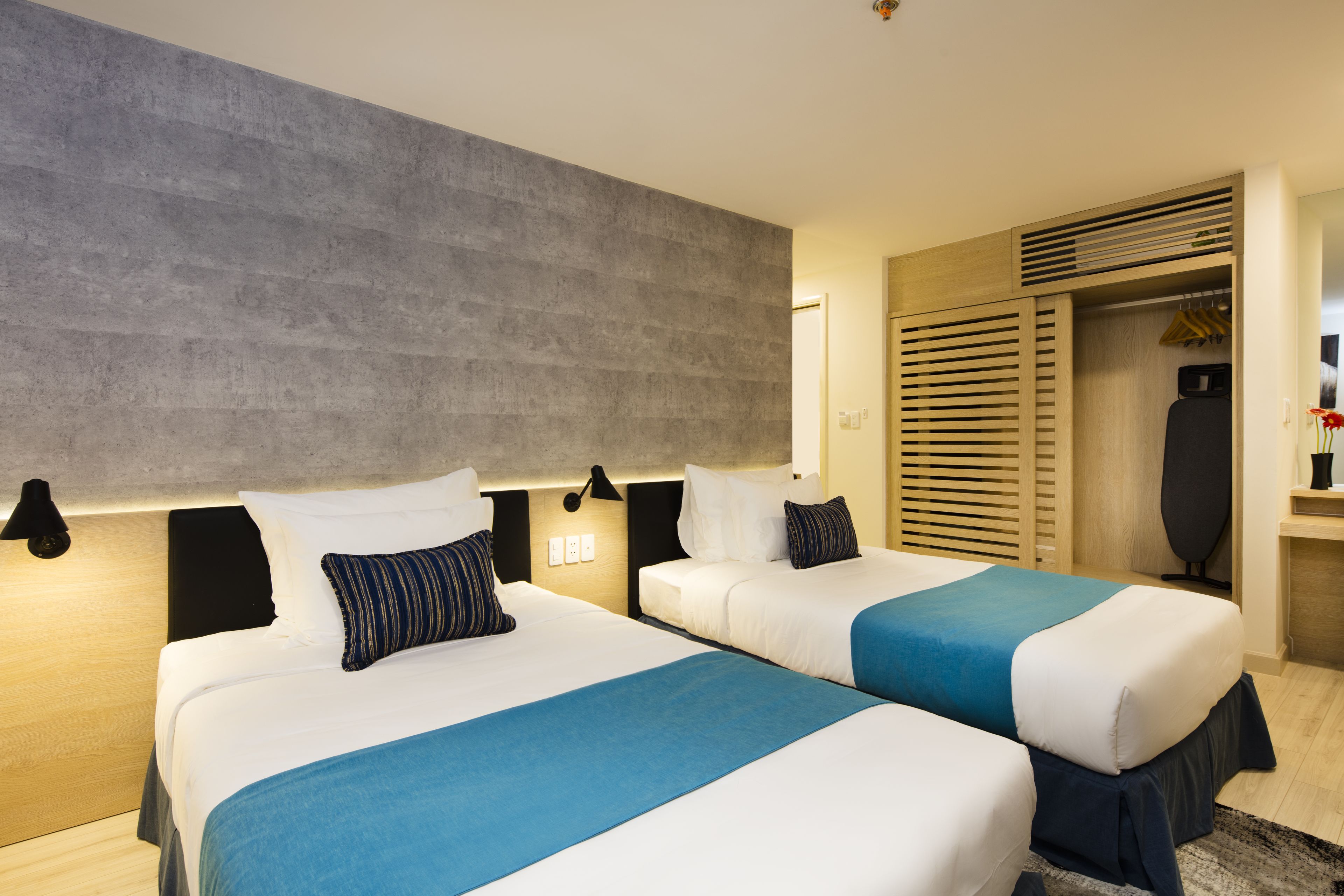Une chambre d’hôtel avec deux lits, un grand mur gris et une armoire en bois où sont suspendus des vêtements.