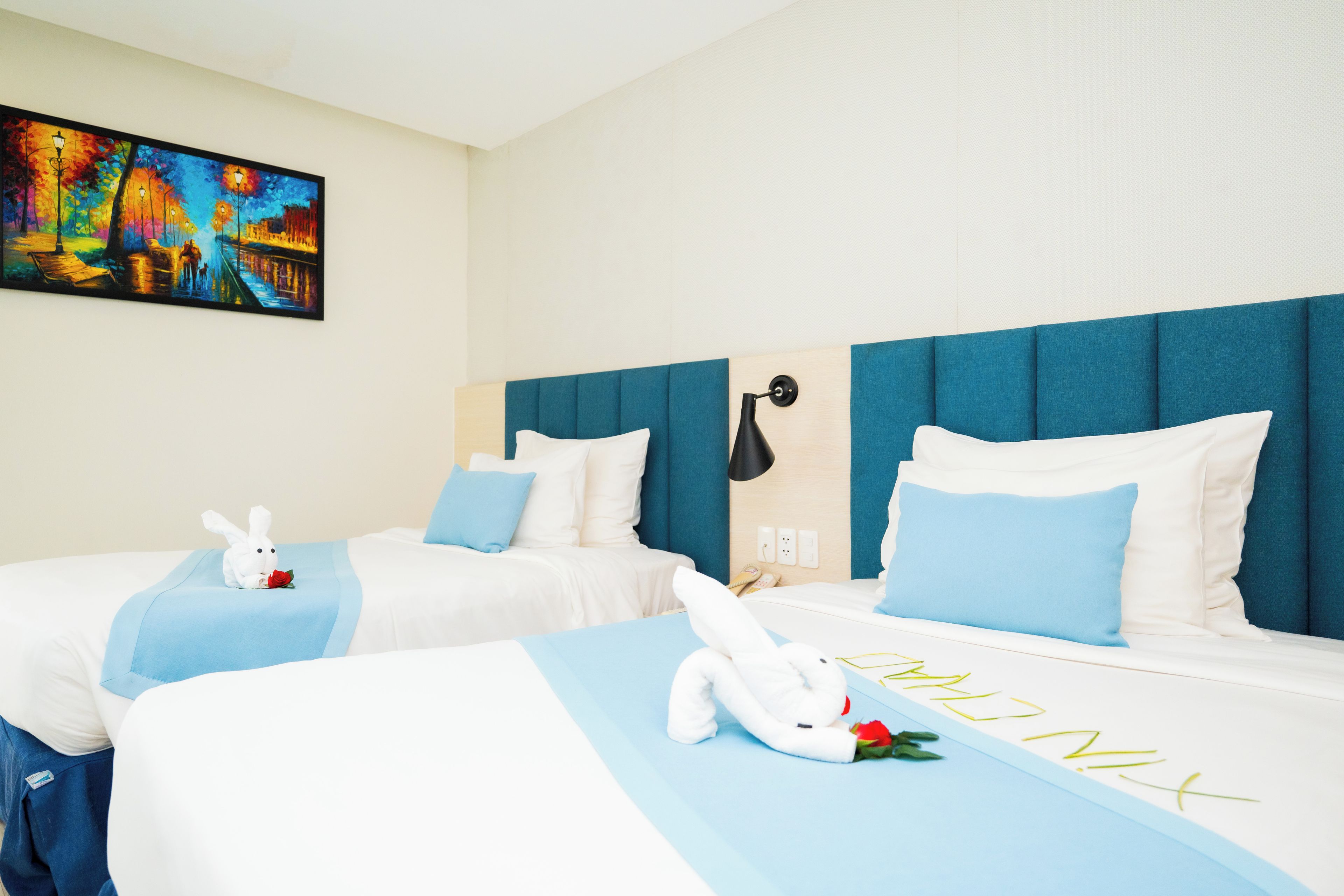 A hotel room with two beds, each with a white comforter and blue accents, a painting on the wall, and a nightstand with a lamp.