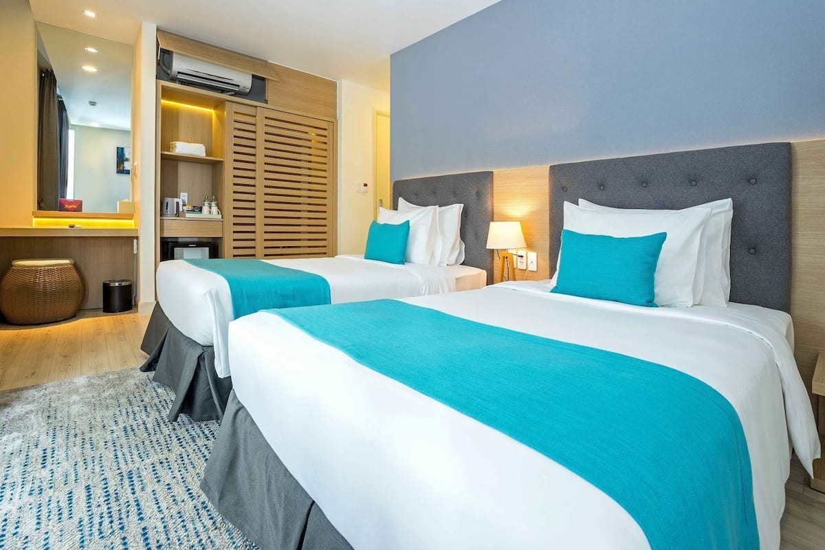 Une chambre d’hôtel avec deux lits, une tête de lit en bois, une table de chevet avec une lampe, un miroir et un petit coin salon avec un fauteuil en osier.