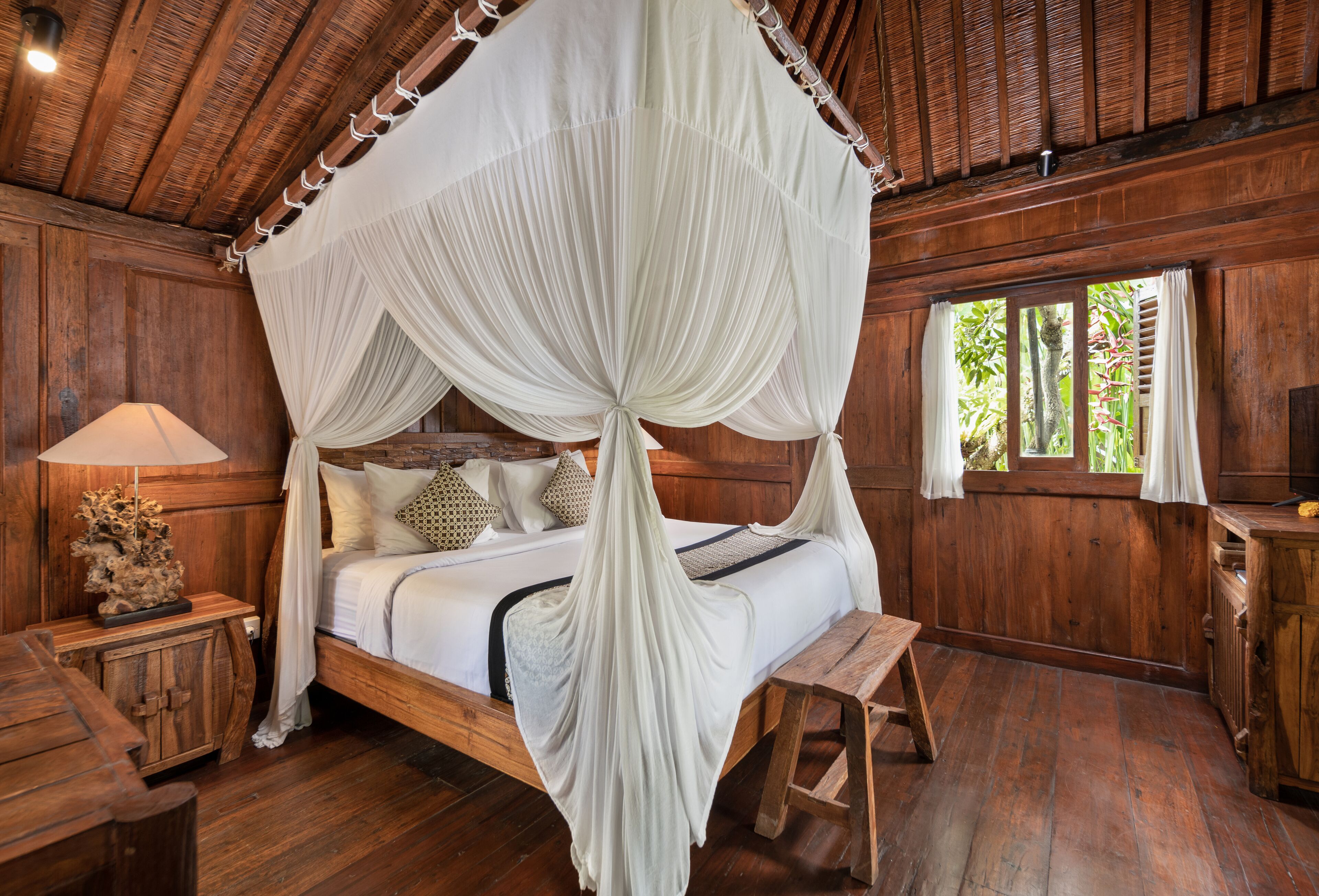 Una cama con dosel de madera con cuatro columnas, una mesita de noche con lámpara y un banco de madera.