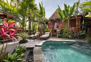 Un área de piscina con terraza de madera, sillas de descanso y plantas tropicales.