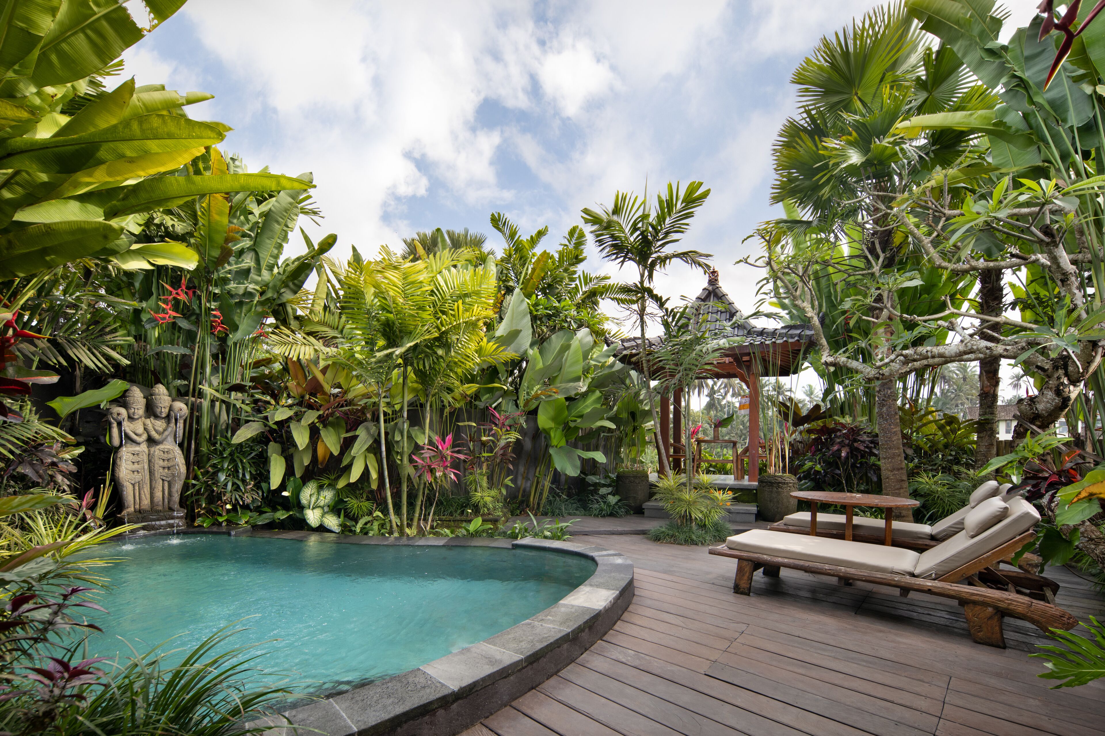 A pool surrounded by lush tropical plants, a wooden deck, and a lounge chair.