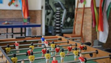 A foosball table in a room with a brick wall, a blue table tennis table, and a staircase.