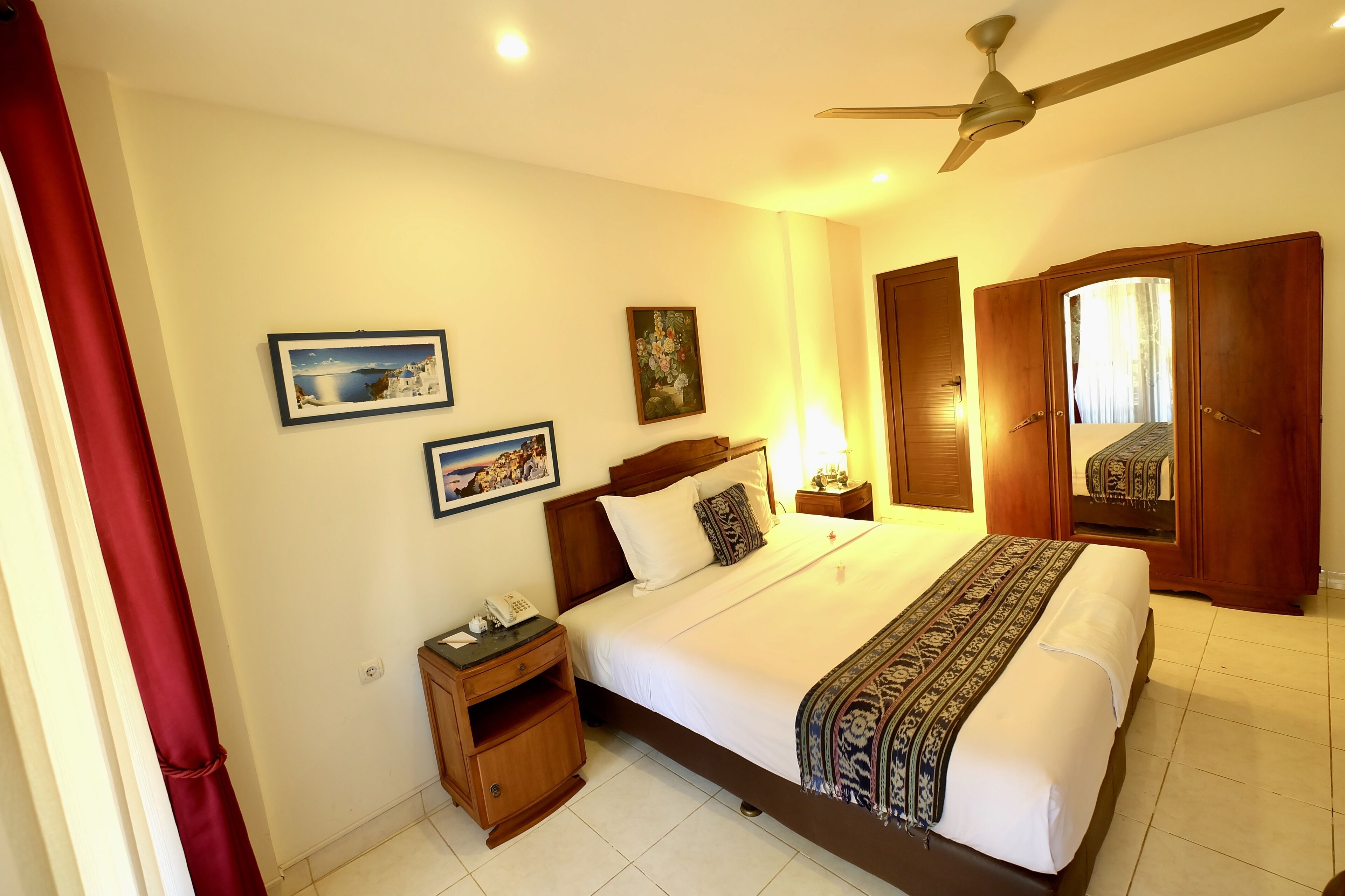 A hotel room with a bed, wooden wardrobe, bedside table, and a ceiling fan.