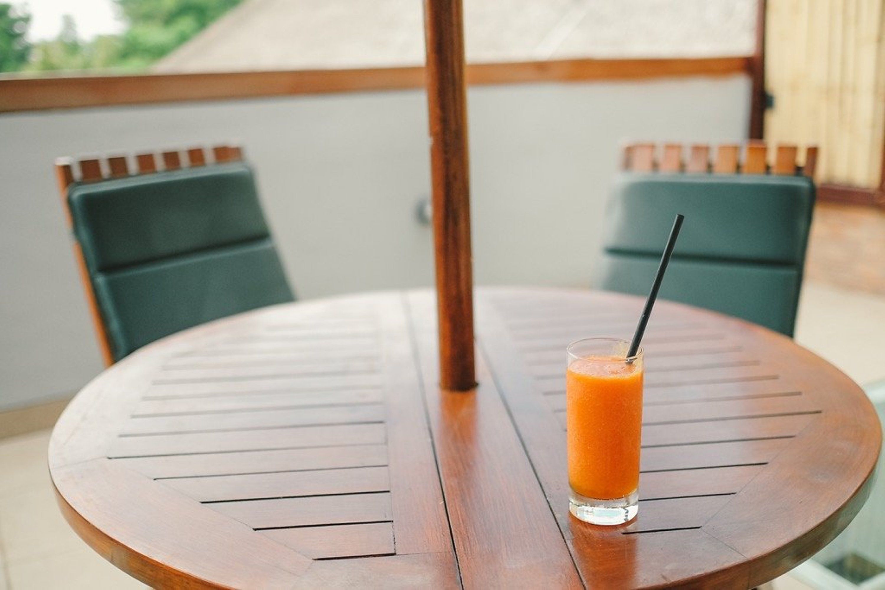 Una mesa de madera redonda con un vaso de jugo de naranja y una pajita, dos sillas verdes y una vista de follaje a través de una ventana.