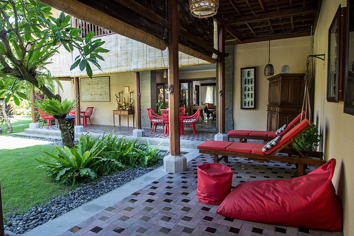 A covered outdoor seating area with red chairs and cushions, a small garden with plants, and a view of the interior through an open doorway.