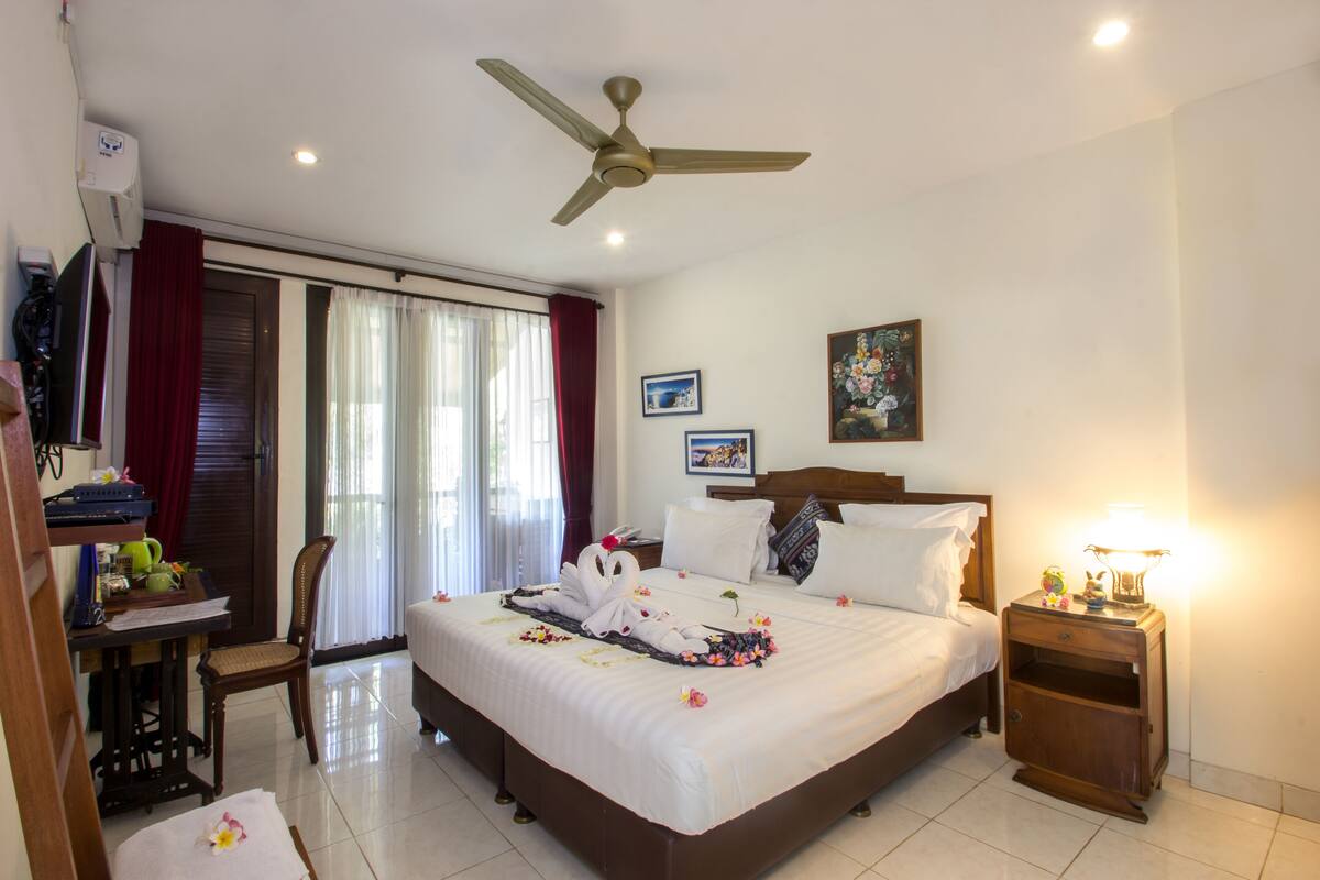 Une chambre d’hôtel comprenant un lit, un bureau, une chaise, une table de chevet, un ventilateur de plafond et une fenêtre avec des rideaux.
