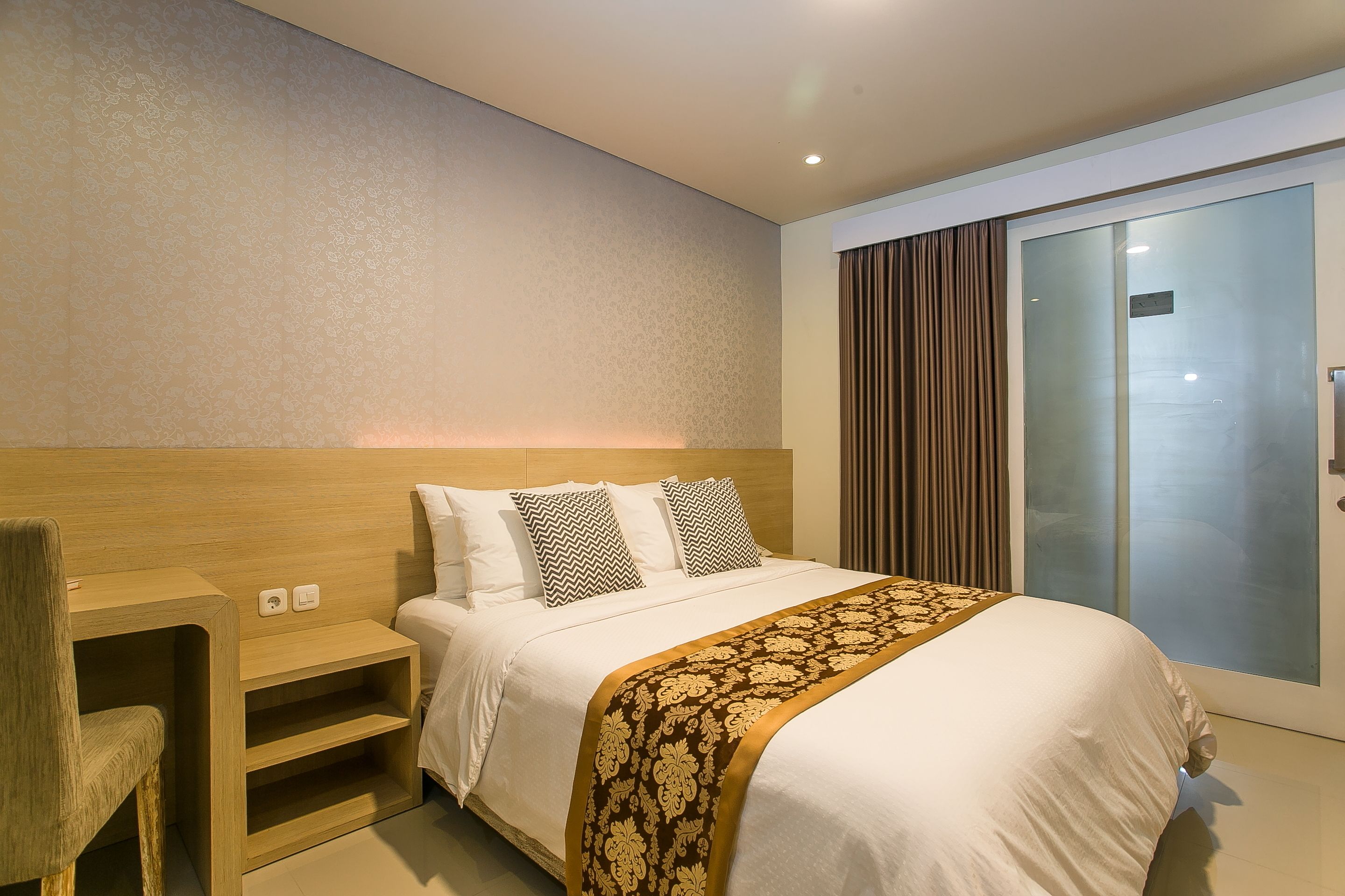 A modern hotel room with a large bed, a wooden nightstand, a built-in shelf, and a sliding glass door leading to a balcony.