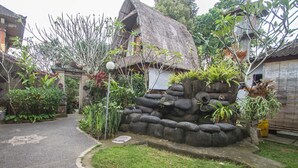 Une maison traditionnelle avec un toit de chaume, entourée d’un jardin bien entretenu où poussent différentes espèces de plantes, et bordée d’un mur de pierre.