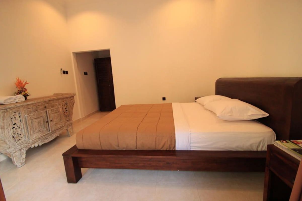 Une chambre à coucher avec un lit en bois, une table de chevet, une commode et une porte donnant sur une autre pièce.