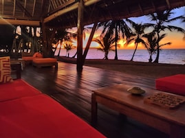 Un salón junto a la playa con piso de madera y asientos acolchados de color rojo, con vista al océano al atardecer.