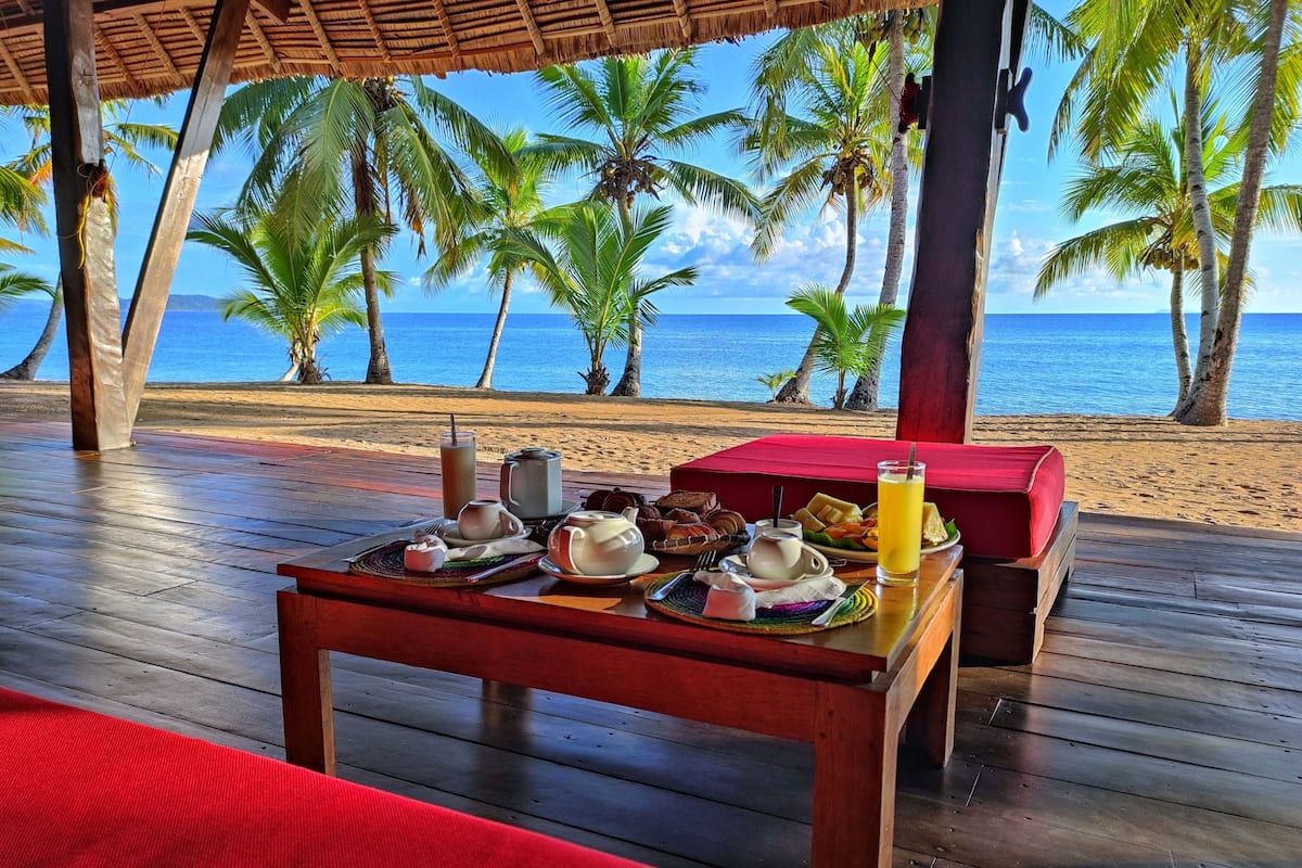 A wooden deck with a table set for breakfast overlooking a beach with palm trees and the ocean.