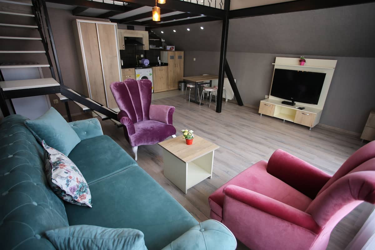 A modern living room with a teal sofa, a red armchair, and a purple armchair. A wooden coffee table with a plant is in the center. A TV is mounted on the wall, and a staircase is visible in the background.