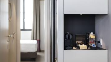 Un pasillo estrecho que da a un dormitorio con cama y ventana. Se ve una kitchenette con cafetera y un refrigerador pequeño.