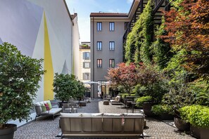 An outdoor seating area with a sofa and chairs, surrounded by potted plants and a building with a yellow stripe. - Savona 18 Suites – Member of Preferred Hotels & Resorts (Milan)