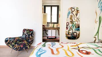 Une pièce comprenant un fauteuil moderne, un lit orné d’un motif abstrait aux couleurs vives, et une salle de bain avec un miroir et des porte-serviettes.