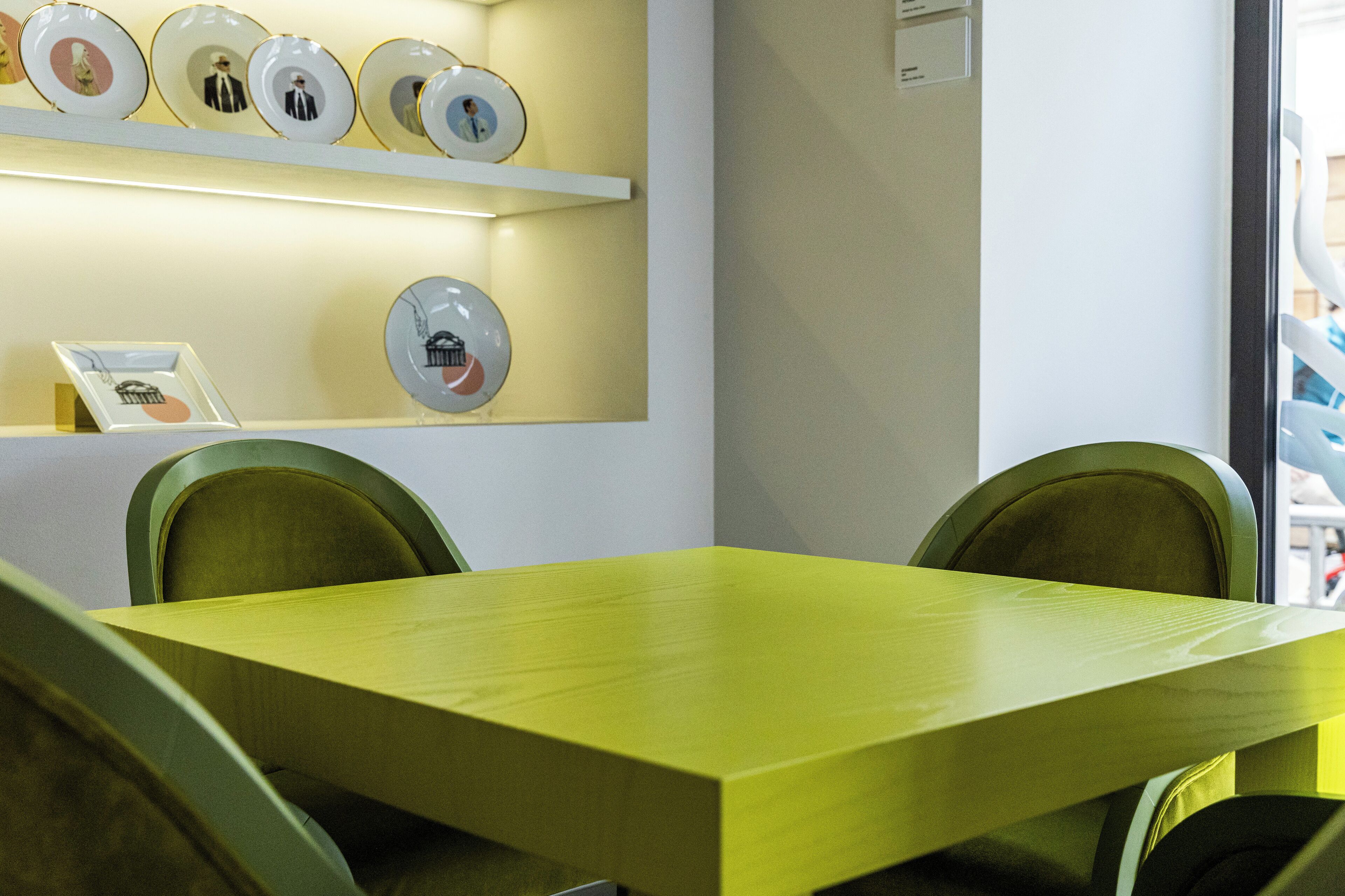 Un salón de conferencias con una mesa amarilla y sillas verdes, un estante montado en la pared con platos decorativos y un cuadro enmarcado.
