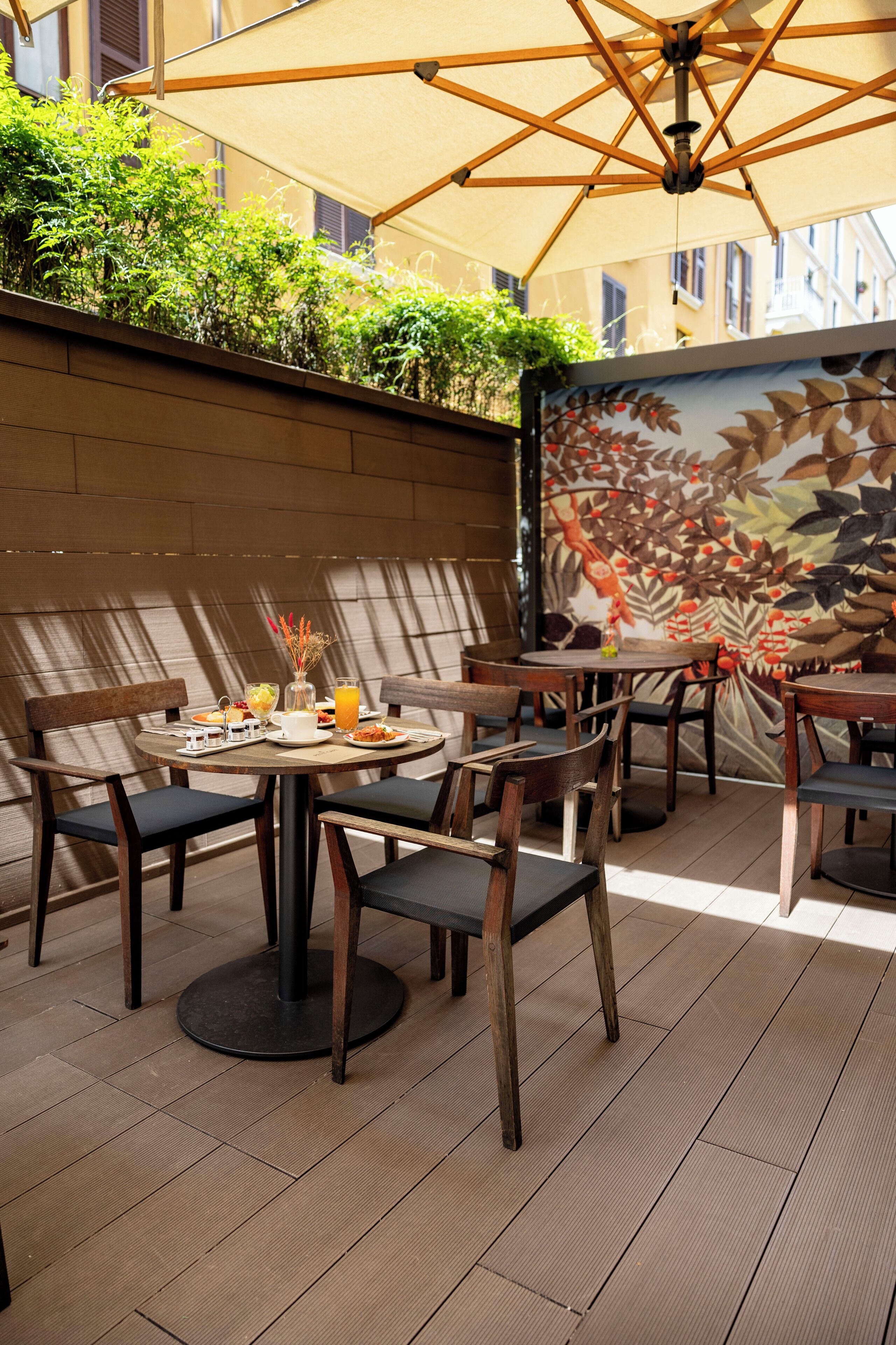 An outdoor dining area with wooden tables and chairs, a mural on the wall, and a large umbrella providing shade.