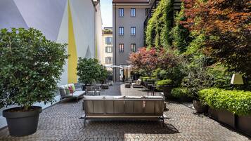An outdoor seating area with a sofa and chairs, surrounded by potted plants and a building with a yellow and white facade.