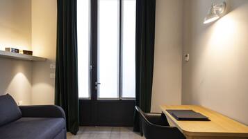 Una habitación pequeña con un sofá, una mesa y una silla. Tiene una ventana con cortinas, una lámpara de pared y un estante con objetos.