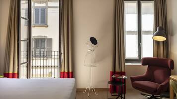 Una habitación de hotel con una cama, un sillón rojo, una lámpara de pie, una mesita y un maniquí con sombrero.