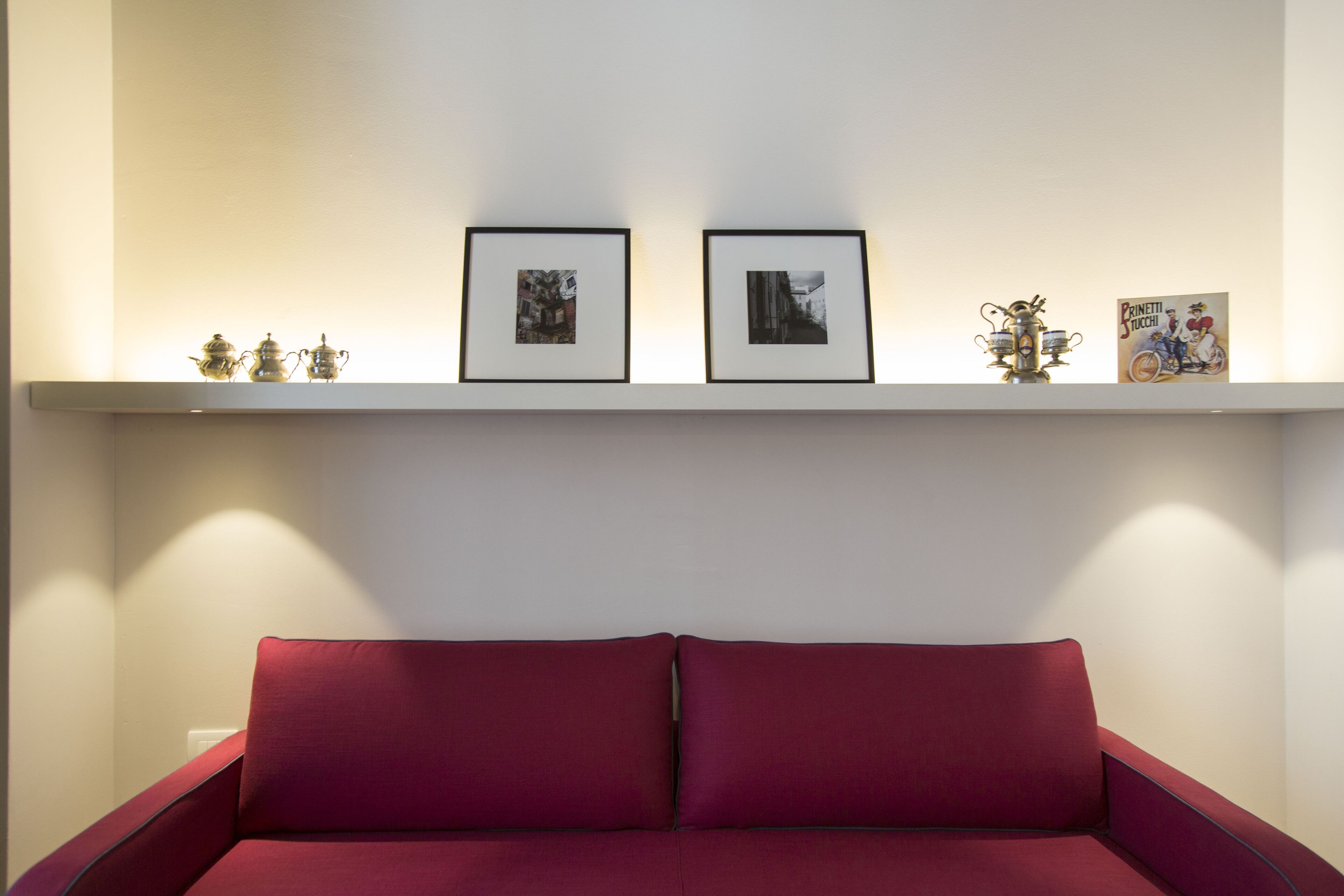 Une pièce avec un canapé rouge, un mur blanc et une étagère ornée de tableaux encadrés et de petits objets décoratifs.